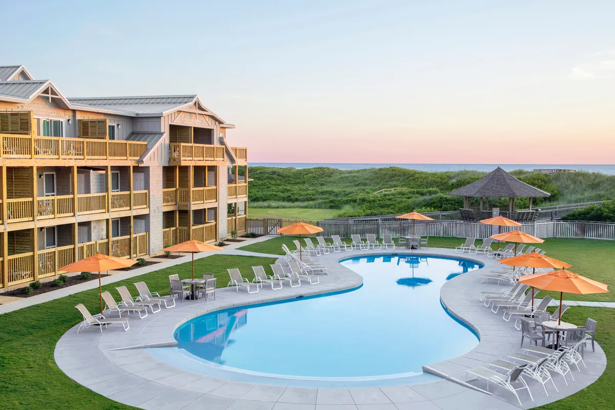This image presents a lively resort pool scene framed by orange umbrellas and lush landscaped lawns, with glimpses of the building’s balconies in the background. Guests are seen swimming, sunbathing, and socializing by the sparkling water, showcasing both relaxation and vibrant resort energy.