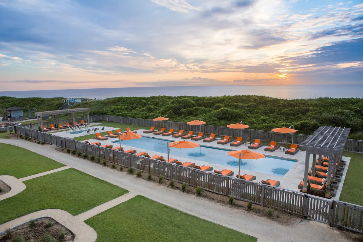 A vibrant outdoor pool area featuring a sparkling swimming pool surrounded by comfortable lounge chairs and bright orange umbrellas, all set against lush, landscaped greenery. The tranquil scene is inviting, with hints of social gatherings and relaxation under the sun.