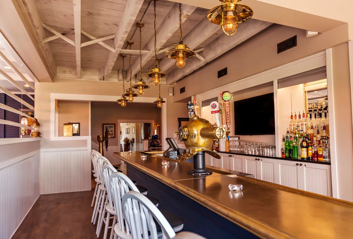 A stylish interior bar with a polished counter, illuminated by elegant hanging lights and a display of bottled drinks. The space feels intimate and welcoming, suggesting a spot to enjoy signature cocktails or a quiet evening drink together after a day by the ocean.