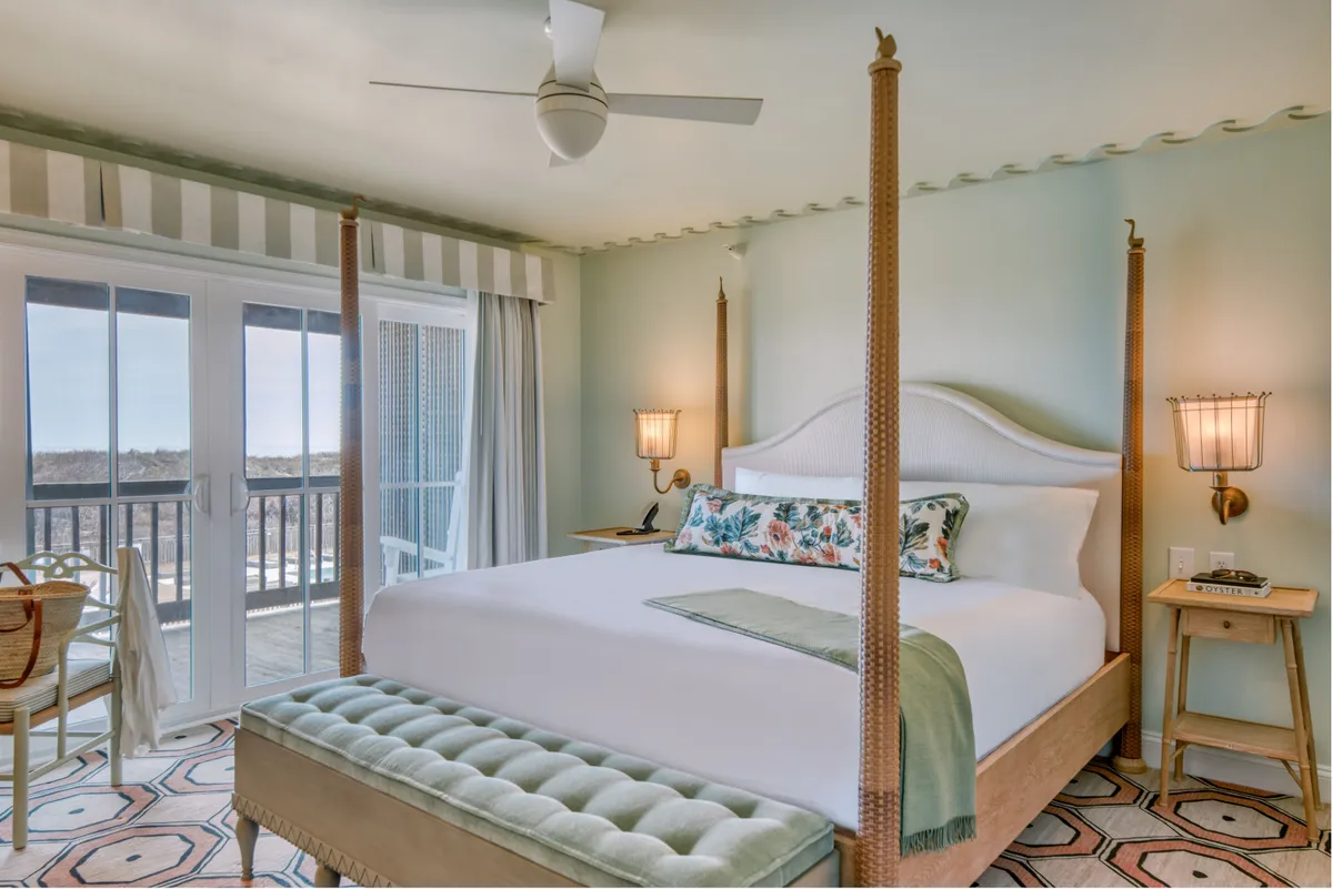 A sunlit king guestroom featuring a four-poster bed with colorful, luxurious bedding. Large windows offer captivating views of the ocean, while a decorative bench sits at the foot of the bed. The muted color palette and soft décor evoke tranquility and comfort, with natural light streaming in to enhance the serene atmosphere.