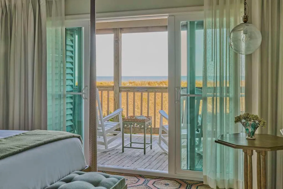 The image reveals an inviting studio interior with open doors leading to a private deck. Two rocking chairs face the Atlantic, perfect for soaking in panoramic ocean views. Soft morning light floods the room, highlighting elegant decor and a decorative table adorned with fresh flowers, setting a serene, romantic mood ideal for couples.