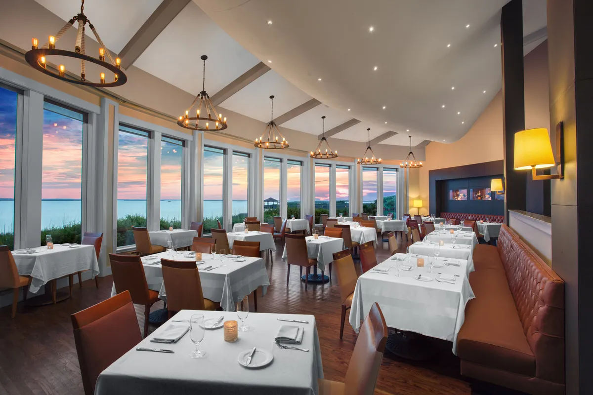 An elegant interior dining room with large windows overlooking a sunset, sophisticated chandeliers, and beautifully set tables. The ambiance is upscale yet inviting, perfect for intimate meals or group celebrations.
