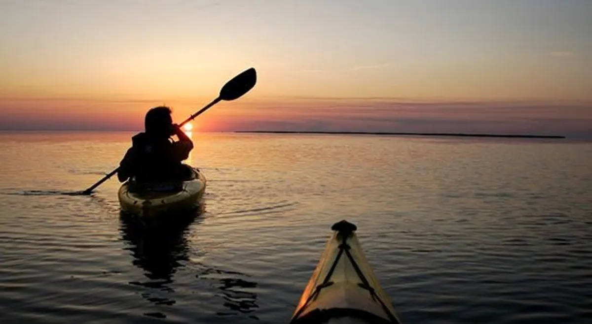 A kayaker glides across calm water at sunset, the sky painted in warm hues and reflecting on the water’s surface. The scene radiates peace, romance, and the allure of memorable sunset adventures. The expansive view puts the focus on the natural beauty and intimate activities available by the ocean.