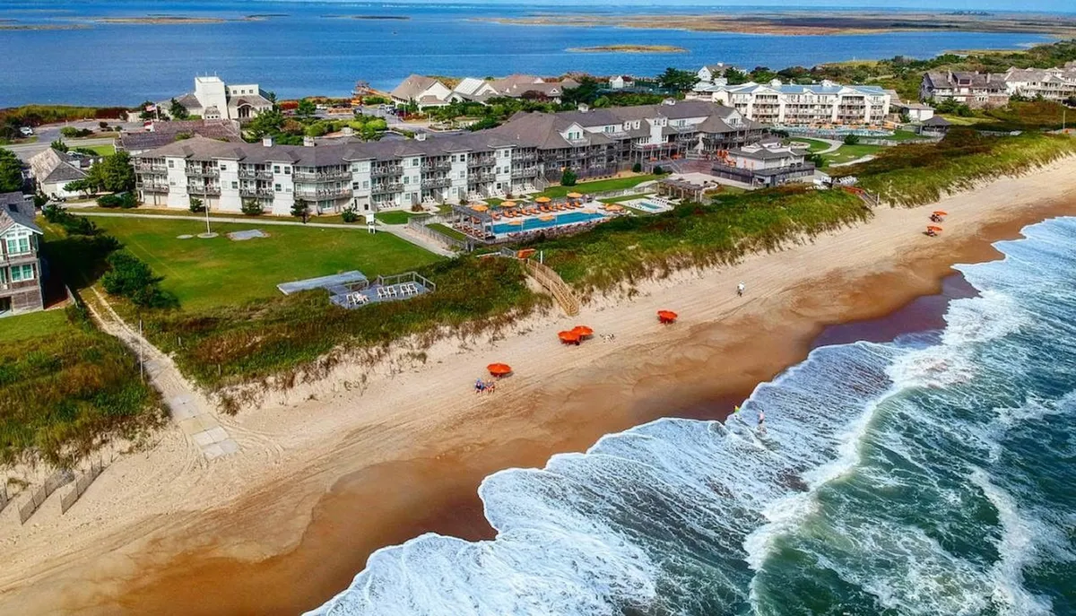 This vibrant exterior image captures the Sanderling Resort's beachfront setting, with colorful umbrellas dotting the sand, a sparkling swimming pool, and the expansive ocean just beyond. Families and groups are visible enjoying sunbathing, swimming, and casual beach sports, creating a lively yet relaxing atmosphere. The overall scene highlights the seamless connection between resort amenities and the natural beauty of the Outer Banks shoreline.