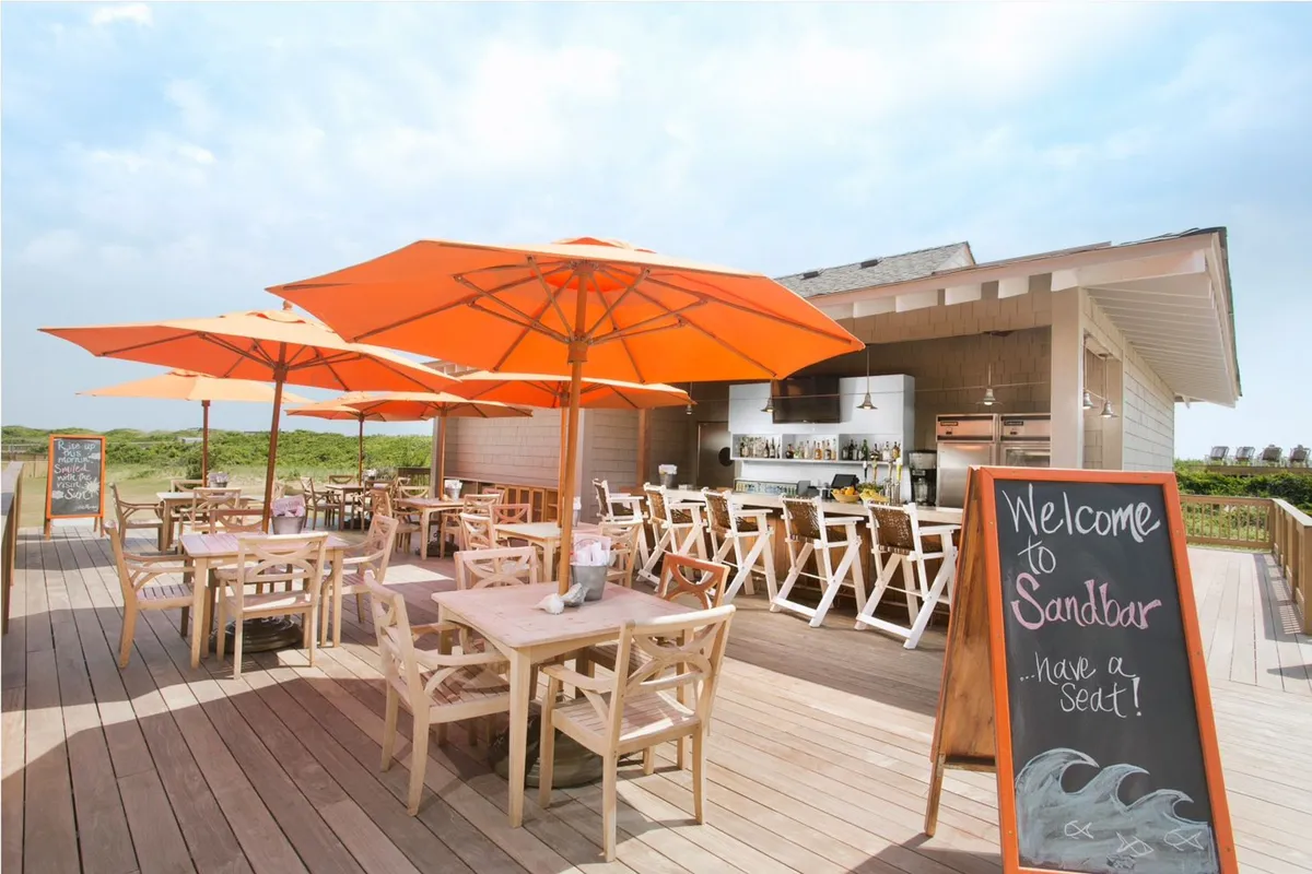 An exterior dining area with wooden tables and chairs under bright orange umbrellas next to a welcoming bar area. The sunny, open-air space hints at a relaxed, friendly atmosphere ideal for enjoying healthy, locally inspired meals or drinks outdoors.
