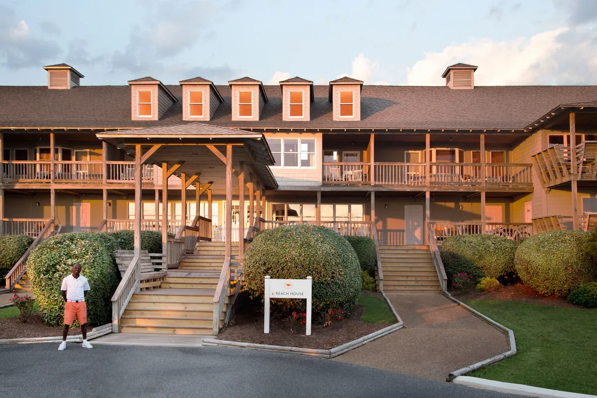 The beach house exterior with stairs leading up to a porch, surrounded by manicured bushes and a sign reading 'A Beach House.' The setting is cheerful, inviting guests to relax on the porch or explore nearby amenities.