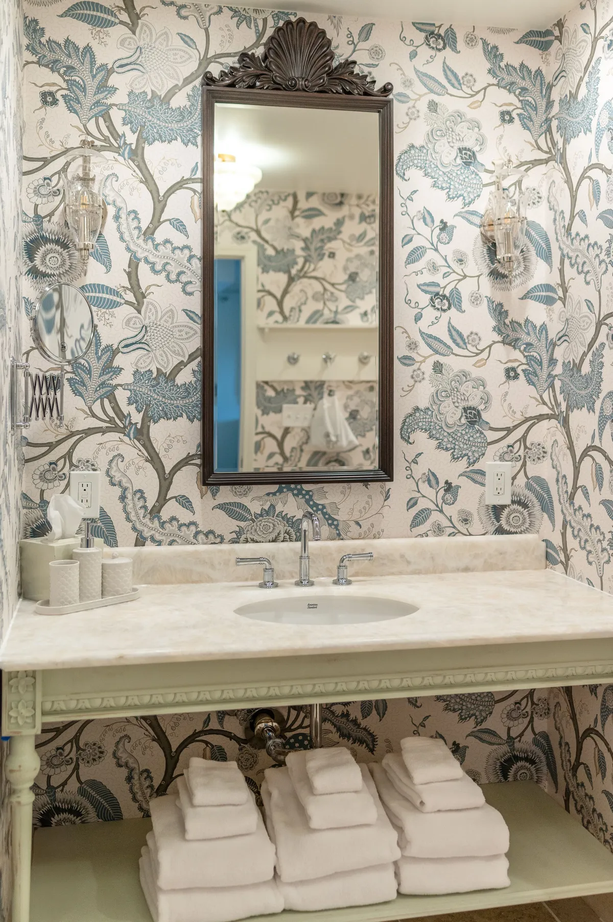 A calming bathroom interior featuring an ornate mirror above a marble countertop, floral wallpaper, and neatly stacked plush towels. The space feels tranquil and inviting, with elegant touches and natural light filtering in, suggesting a place designed for rejuvenation and self-care.