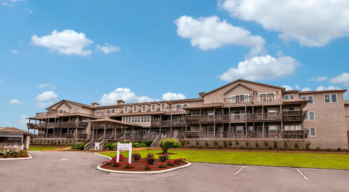 The image shows the building's facade with several inviting balconies overlooking landscaped gardens. The balconies, some furnished and visible, hint at private outdoor spaces perfect for relaxation. The tranquil setting is enhanced by the soft sunlight, suggesting both morning and evening enjoyment from the balcony. The landscaping and overall ambiance evoke a sense of peaceful seclusion, aligning with the narrative of a private retreat.
