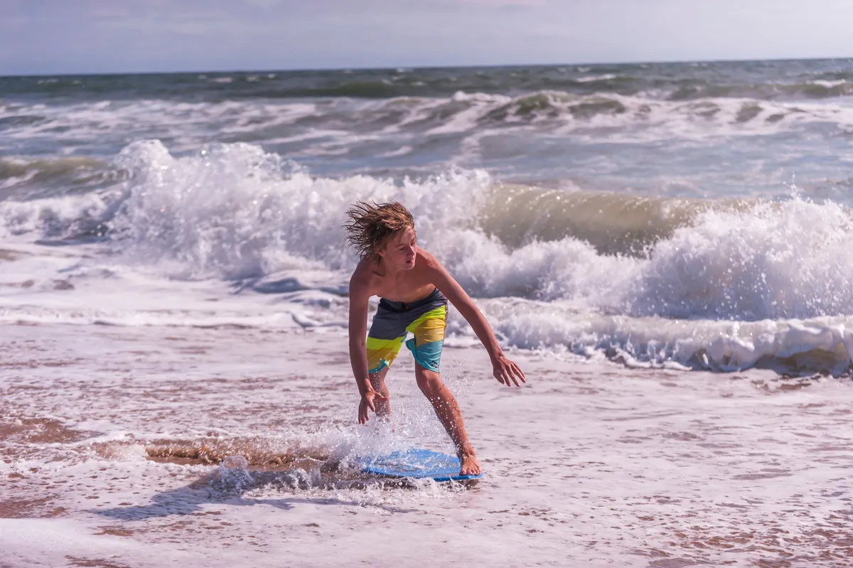 This exterior image features a boy riding a boogie board as waves crash onto the sunlit shore, encapsulating the thrill of water sports at OBX. The sunlight glistens across the water, with hints of sandcastles and other guests enjoying the beach in the background. The mood is playful and energetic, underlining the sense of outdoor adventure.