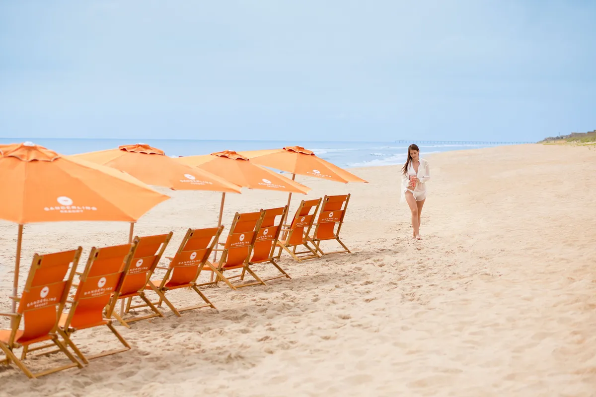 The image captures an inviting beach scene with sun loungers and umbrellas set up on the sand, facing the rolling Atlantic waves. A figure walks along the shoreline, suggesting solitude and reflection. The view emphasizes the expanse of beach and unobstructed ocean, echoing the panoramic vistas available from the balcony above.