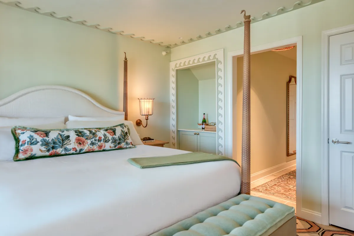 A calm, inviting interior with a neatly made bed adorned with a floral pillow and a green accent blanket. The room includes a decorative light fixture, a cozy seating area, and a mirror, all bathed in natural light from large windows. The setting radiates a sense of peace and comfort, perfect for relaxation or enjoying a quiet morning.