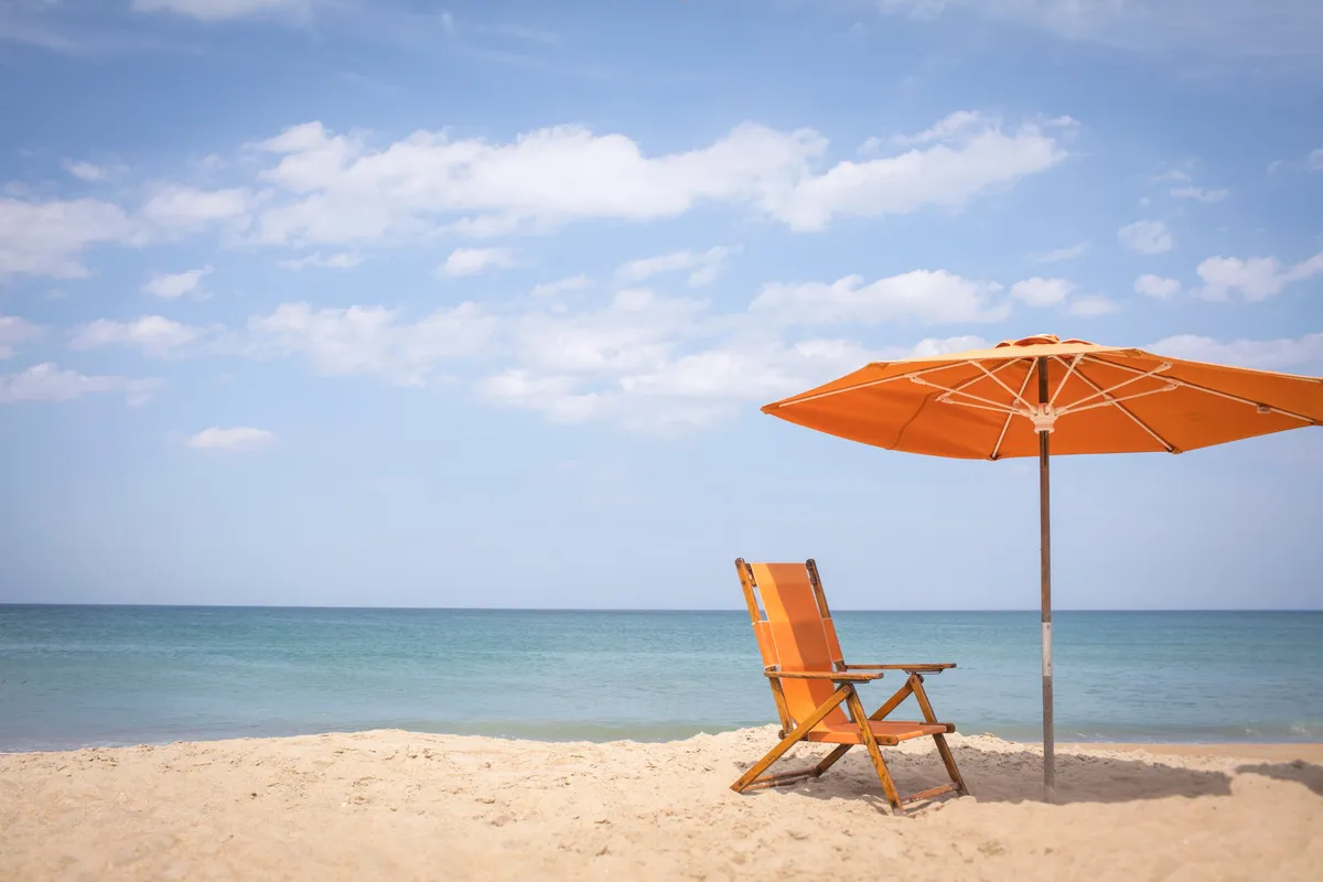 A serene stretch of sandy beach with a single lounge chair and a colorful umbrella facing the sparkling Atlantic Ocean. Gentle waves roll onto the shore, creating a tranquil atmosphere perfect for relaxing, sunbathing, or sharing a quiet moment. The scene captures the peacefulness and romance of a private seaside escape.
